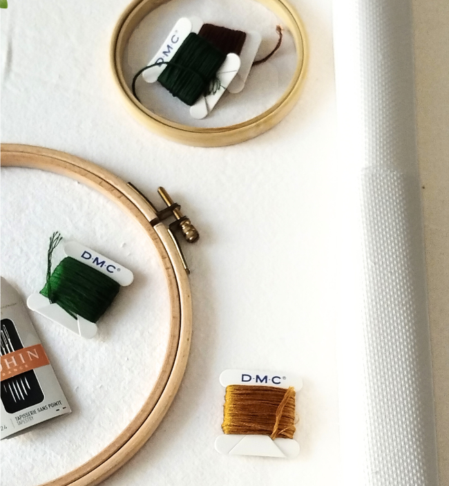 Matériel de broderie et point de croix avec tambour en bois, échevettes de fils DMC, aiguilles et rouleau de toile Aïda blanche sur fond clair.