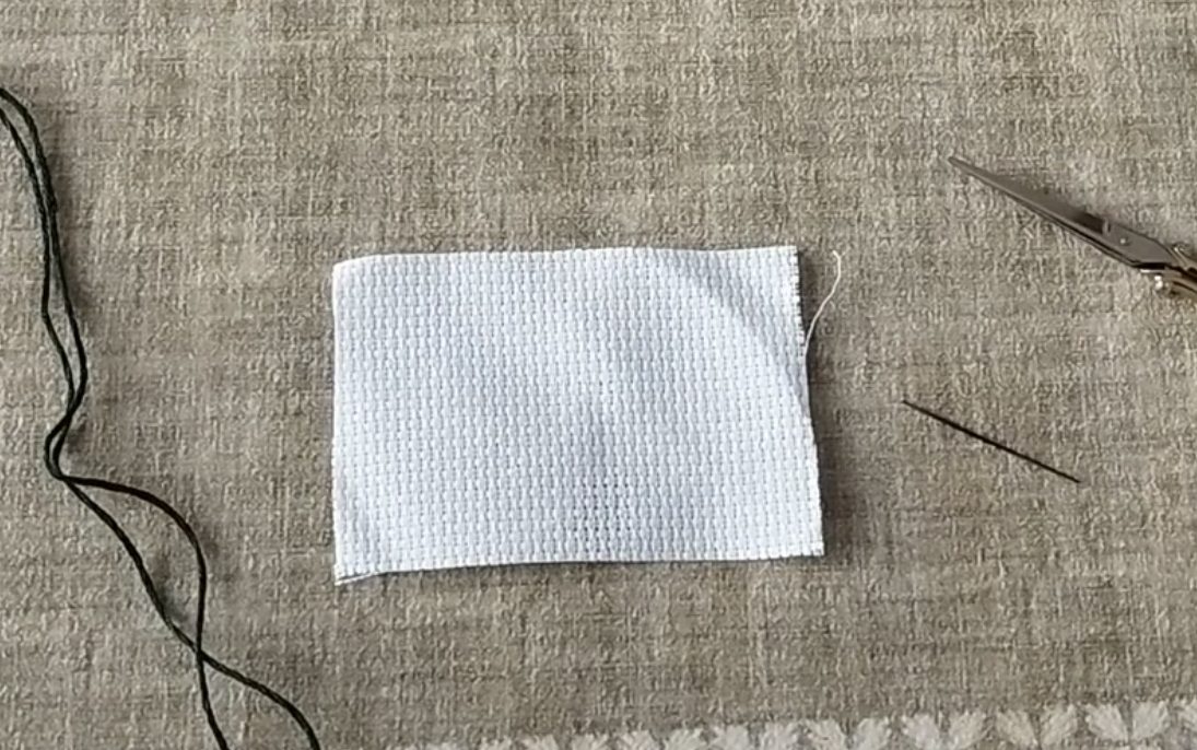 Sur une nappe sont posées : un morceau de toile Aida, une aiguille, une paire de ciseaux et un fil